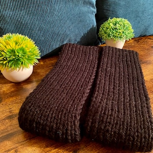 May include: Two black knitted scarves folded on a wooden surface. The scarves are made of a thick, textured yarn.