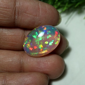 May include: An oval-shaped cabochon opal gemstone featuring a translucent, milky white base with vibrant, multi-colored flashes of green, red, and orange fire throughout the stone, displaying a high-domed surface and a polished, smooth finish.