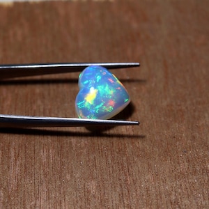 May include: A heart-shaped opal gemstone held by metal tweezers. The stone features a translucent white base with vibrant flashes of green, blue, and yellow light reflecting from within its polished, smooth, and rounded surface structure.