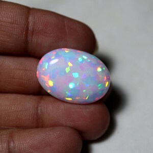 May include: An oval-shaped cabochon gemstone with a milky white base and vibrant, multi-colored iridescent flashes of blue, green, yellow, and pink. The stone features a smooth, polished surface that reflects light, creating a play of color across its rounded, symmetrical form.