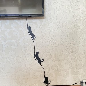 Könnte beinhalten: Drei schwarze Katzensilhouetten sind an einem schwarzen Kabel befestigt, das an einer weißen Wand mit Blumenmuster herunterläuft. Das Kabel ist an einer Steckdose am unteren Ende der Wand angeschlossen.