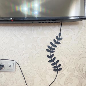 Può includere: Un organizer per cavi nero a forma di vite con foglie è attaccato a un muro. L'organizer viene utilizzato per nascondere un cavo nero proveniente da un televisore. Sul lato sinistro dell'immagine è visibile una presa a muro.