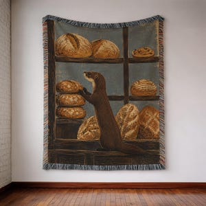 May include: A woven throw blanket featuring a dark brown ferret standing on its hind legs in front of wooden shelves filled with various round and oval loaves of bread. The blanket has a fringed edge with multi-colored threads along the sides.