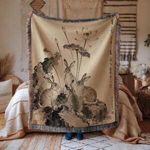 May include: A woven throw blanket featuring a beige background with a black ink-style illustration of three rabbits among lotus flowers and leaves. The blanket has a fringed edge and includes vertical Asian calligraphy text and red stamps.