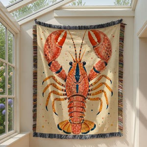 May include: A woven throw blanket featuring a large, detailed illustration of a lobster in shades of orange, red, and blue against a cream-colored background. The blanket has a fringed edge with multicolored threads along the vertical sides.