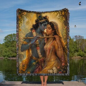 May include: A woven throw blanket featuring a detailed illustration of the deities Krishna and Radha. Krishna has blue skin and holds a golden flute, while Radha wears a yellow sari and veil. They are adorned with floral garlands and jewelry.