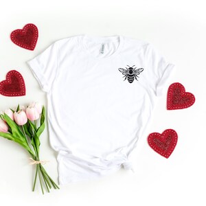 May include: White t-shirt with a black bee graphic on the front. The shirt is surrounded by red hearts and pink tulips.