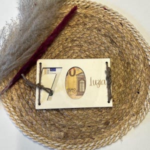 May include: A wooden gift card holder with a cutout for a Euro bill, displaying the number 70. The word "Luzia" is written in cursive. The holder is tied with twine and rests on a woven mat.