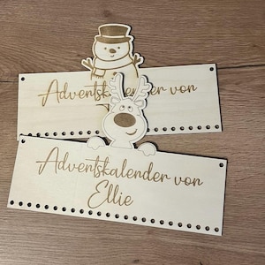 May include: Two wooden Advent calendar signs with laser-cut text and decorative cutouts of a snowman and a reindeer. The signs are light-colored with a series of small holes along the bottom edge.