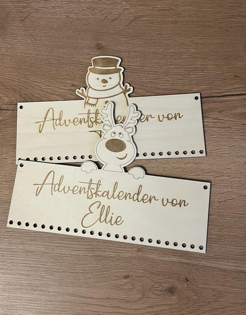 May include: Two wooden Advent calendar signs with laser-cut text and decorative cutouts of a snowman and a reindeer. The signs are light-colored with a series of small holes along the bottom edge.