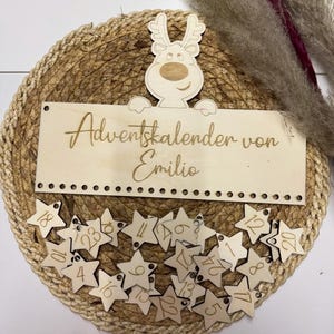 May include: A wooden Advent calendar with star-shaped ornaments numbered 1-24. The calendar features a reindeer design and the text "Adventskalender von Emilio". The calendar is set on a woven mat.