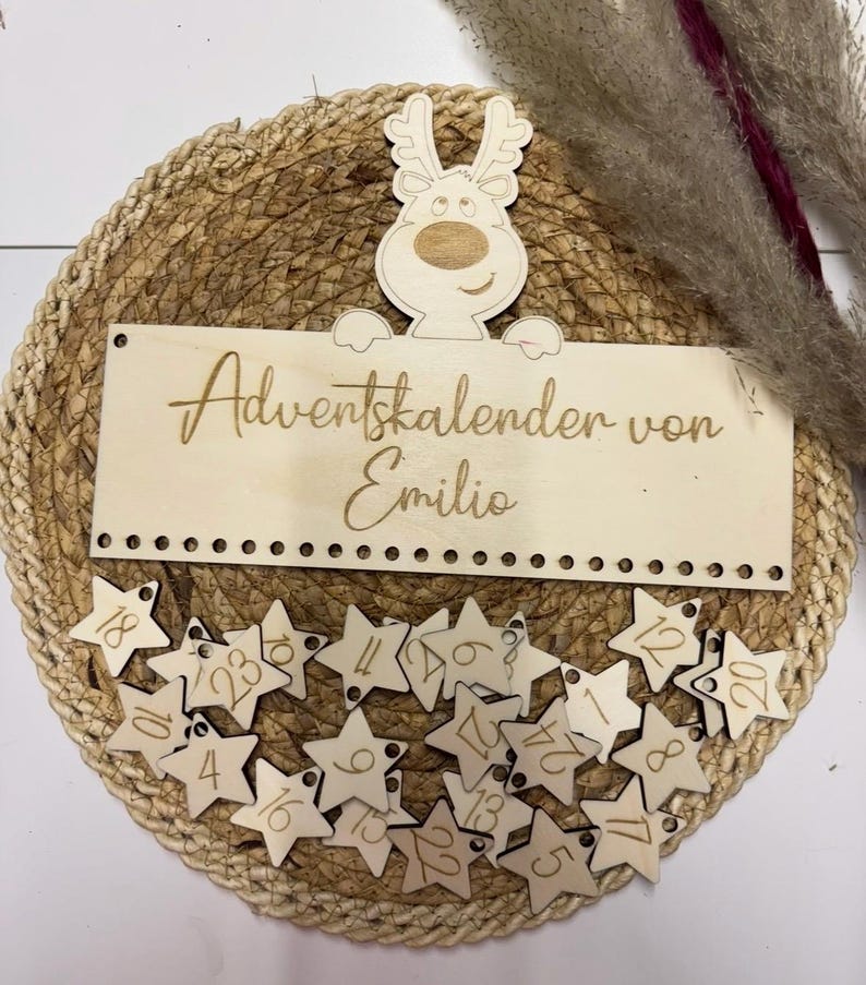 May include: A wooden Advent calendar with star-shaped ornaments numbered 1-24. The calendar features a reindeer design and the text "Adventskalender von Emilio". The calendar is set on a woven mat.