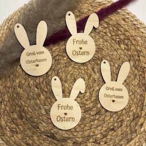 May include: Three wooden bunny-shaped ornaments with different Easter greetings. The first ornament says "Gruss vom Osterhasen" which translates to "Greetings from the Easter Bunny". The second ornament says "Frohe Ostern" which translates to "Happy Easter". The third ornament says "Gruss vom Osterhasen" which translates to "Greetings from the Easter Bunny".
