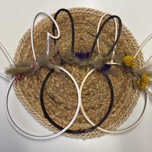 May include: Decorative Easter bunny ear wall art. The ears are made of wood and come in white, black, and natural wood tones. They are adorned with dried flowers in shades of yellow, purple, and pink, set against a woven background.