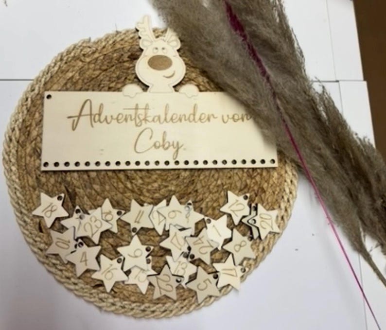 May include: A round Advent calendar with a woven jute base. A wooden sign with the text "Adventskalender von Coby" is at the top, and star-shaped tags numbered 1-24 are below. A wooden reindeer decorates the top.
