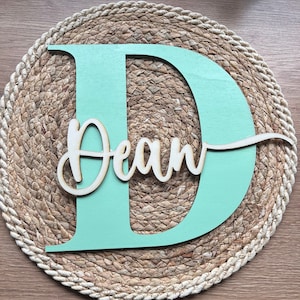May include: A light green wooden letter D with the name "Dean" written in cursive on a natural woven round mat.
