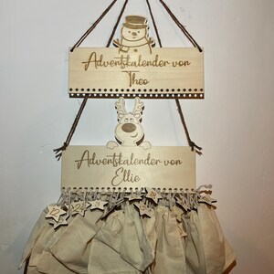 May include: Wooden Advent calendar with two signs, one with a snowman and the text "Adventskalender von Theo", and the other with a reindeer and the text "Adventskalender von Ellie". Below are 24 beige fabric bags with star-shaped tags.