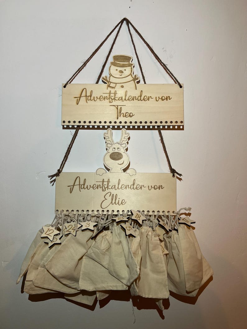 May include: Wooden Advent calendar with two signs, one with a snowman and the text "Adventskalender von Theo", and the other with a reindeer and the text "Adventskalender von Ellie". Below are 24 beige fabric bags with star-shaped tags.