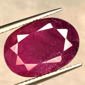 Può includere: Un rubino di forma ovale, tenuto da una pinzetta. La gemma mostra una tonalità rosso intenso con sfaccettature interne che creano un motivo a forma di stella. La superficie lucida riflette la luce, mostrando la sua chiarezza e brillantezza. Una vista ravvicinata evidenzia il colore ricco e il design intricato della gemma.