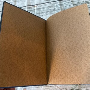 May include: An open notebook with a dark blue cover and brown pages. The pages have a textured appearance. The notebook is lying open on a light-colored, weathered wooden surface.