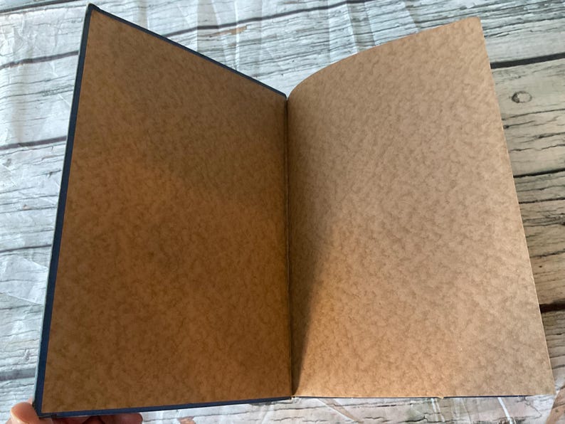 May include: An open notebook with a dark blue cover and brown pages. The pages have a textured appearance. The notebook is lying open on a light-colored, weathered wooden surface.
