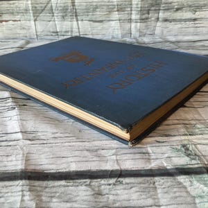 May include: A vintage book with a blue cover and gold lettering. The title "History of the 25th Infantry" is visible. The book's pages have a golden hue, and the cover shows signs of wear, suggesting its age.