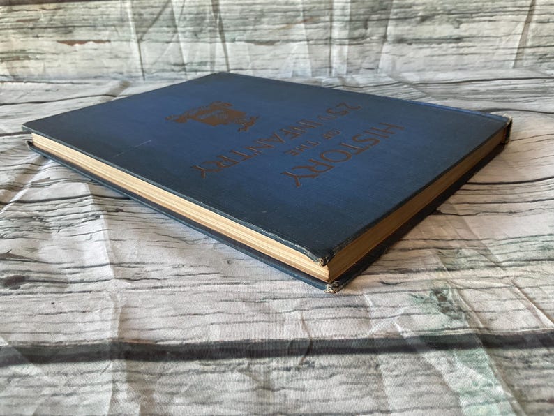 May include: A vintage book with a blue cover and gold lettering. The title "History of the 25th Infantry" is visible. The book's pages have a golden hue, and the cover shows signs of wear, suggesting its age.