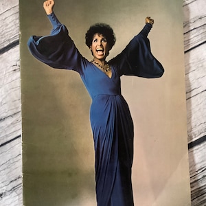 May include: A woman in a blue dress with long sleeves and a plunging neckline is standing with her arms raised in the air. She is smiling and appears to be singing.