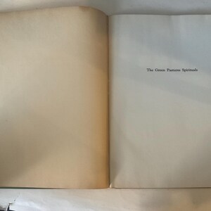 Op de afbeelding: Open boek met verouderde, gebroken witte pagina's. De tekst "The Green Pastures Spirituals" is zwart gedrukt, gecentreerd op de rechterpagina. De linkerpagina heeft een warme, bruine tint.