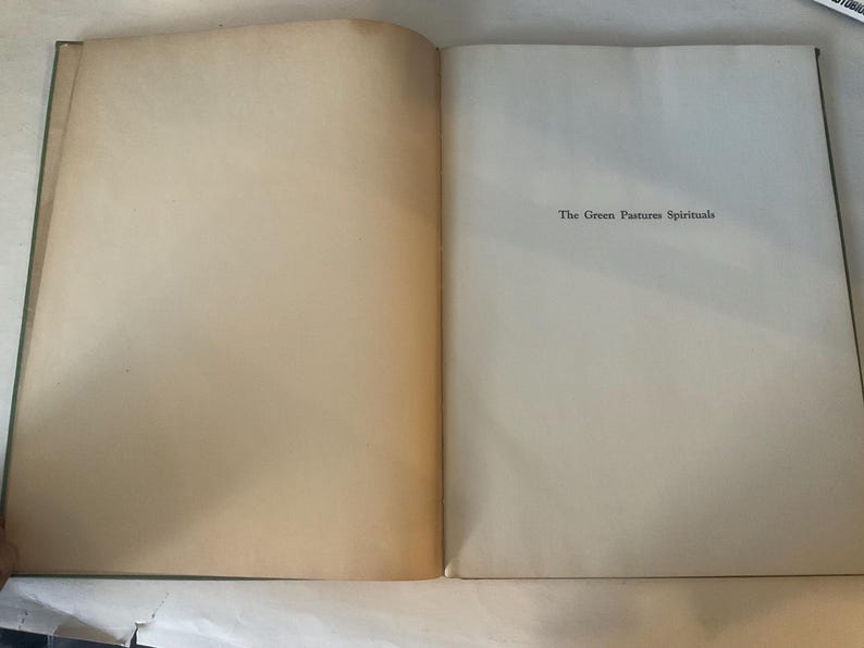 Op de afbeelding: Open boek met verouderde, gebroken witte pagina's. De tekst "The Green Pastures Spirituals" is zwart gedrukt, gecentreerd op de rechterpagina. De linkerpagina heeft een warme, bruine tint.