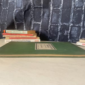 Op de afbeelding: Een stapel boeken met titels als "Mastering French Vocabulary" en "The Wisdom of Confucius". Een groot groen boek staat op de voorgrond, met een label in het midden. De achtergrond is een donkere, getextureerde muur.