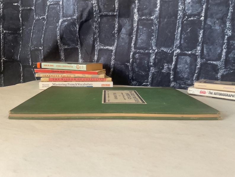 Op de afbeelding: Een stapel boeken met titels als "Mastering French Vocabulary" en "The Wisdom of Confucius". Een groot groen boek staat op de voorgrond, met een label in het midden. De achtergrond is een donkere, getextureerde muur.