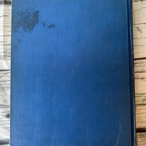 May include: A vintage book with a textured, cobalt blue cover. The cover shows some wear and discoloration. The book's spine is visible on the right side. The background is a light-colored, wooden surface.