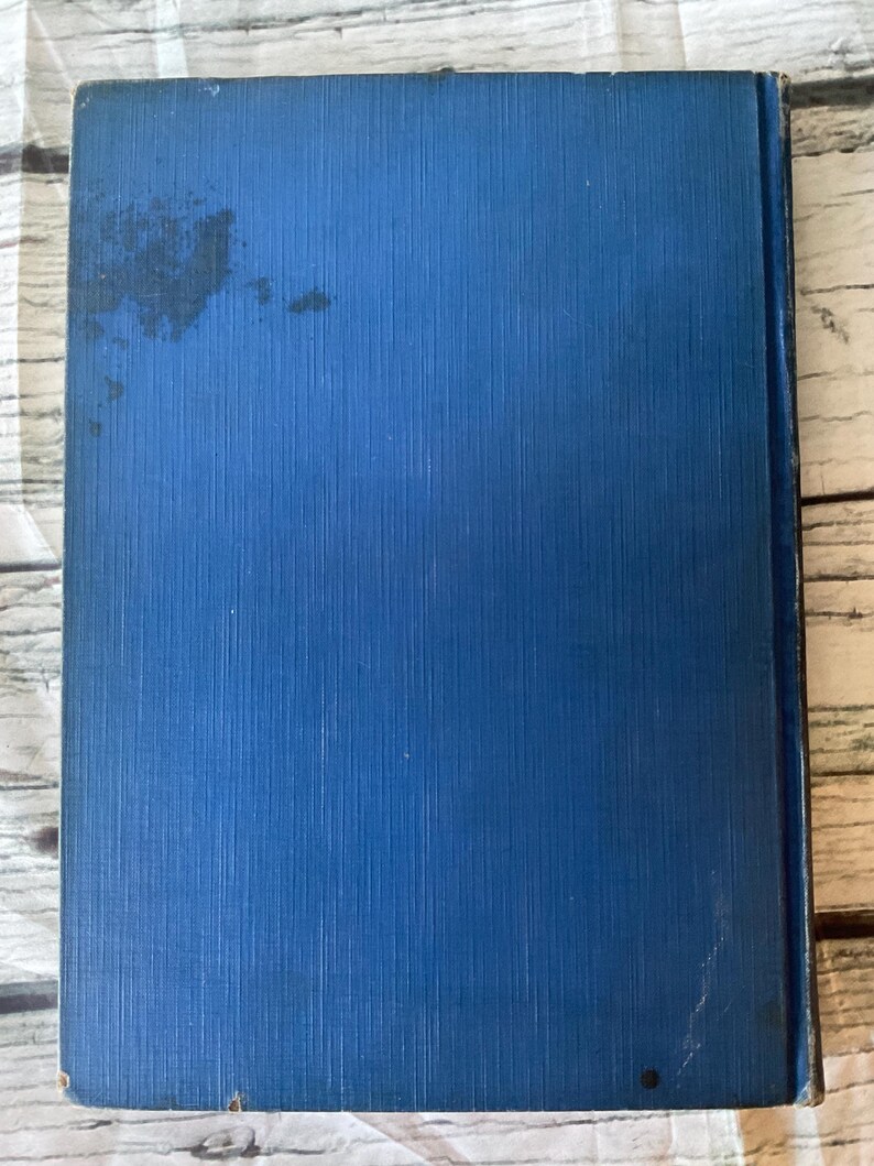 May include: A vintage book with a textured, cobalt blue cover. The cover shows some wear and discoloration. The book's spine is visible on the right side. The background is a light-colored, wooden surface.