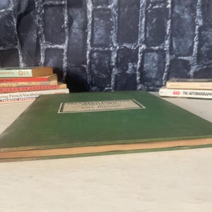 Op de afbeelding: Een vintage groen boek met een rechthoekig label op de kaft. Het label heeft zwarte tekst op een witte achtergrond. Op de achtergrond zijn verschillende andere boeken opgestapeld.