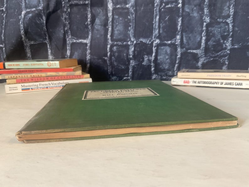 Op de afbeelding: Een vintage groen boek met een rechthoekig label op de kaft. Het label heeft zwarte tekst op een witte achtergrond. Op de achtergrond zijn verschillende andere boeken opgestapeld.