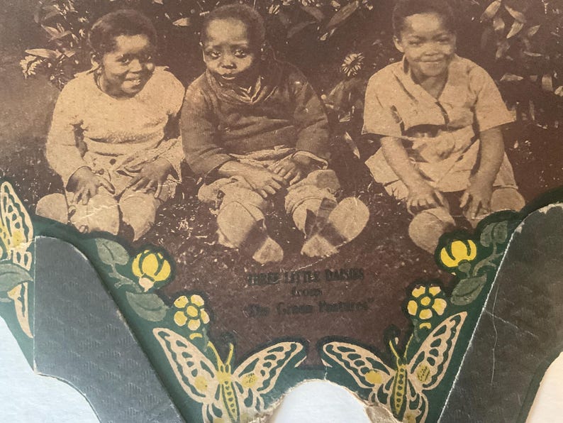 Op de afbeelding: Sepia-getinte afbeelding van drie kinderen die op gras zitten. De afbeelding is omlijst door een decoratieve rand met vlinders en bloemmotieven. De tekst luidt "Three Little Babies from The Green Pastures."