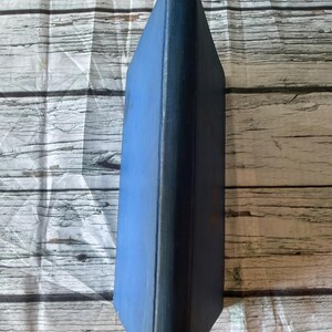 May include: A vintage book with a blue cover and spine. The book's edges are worn, showing signs of age. The cover is a solid blue, while the spine is a darker shade. The book is set against a light-colored, wood-grain background.