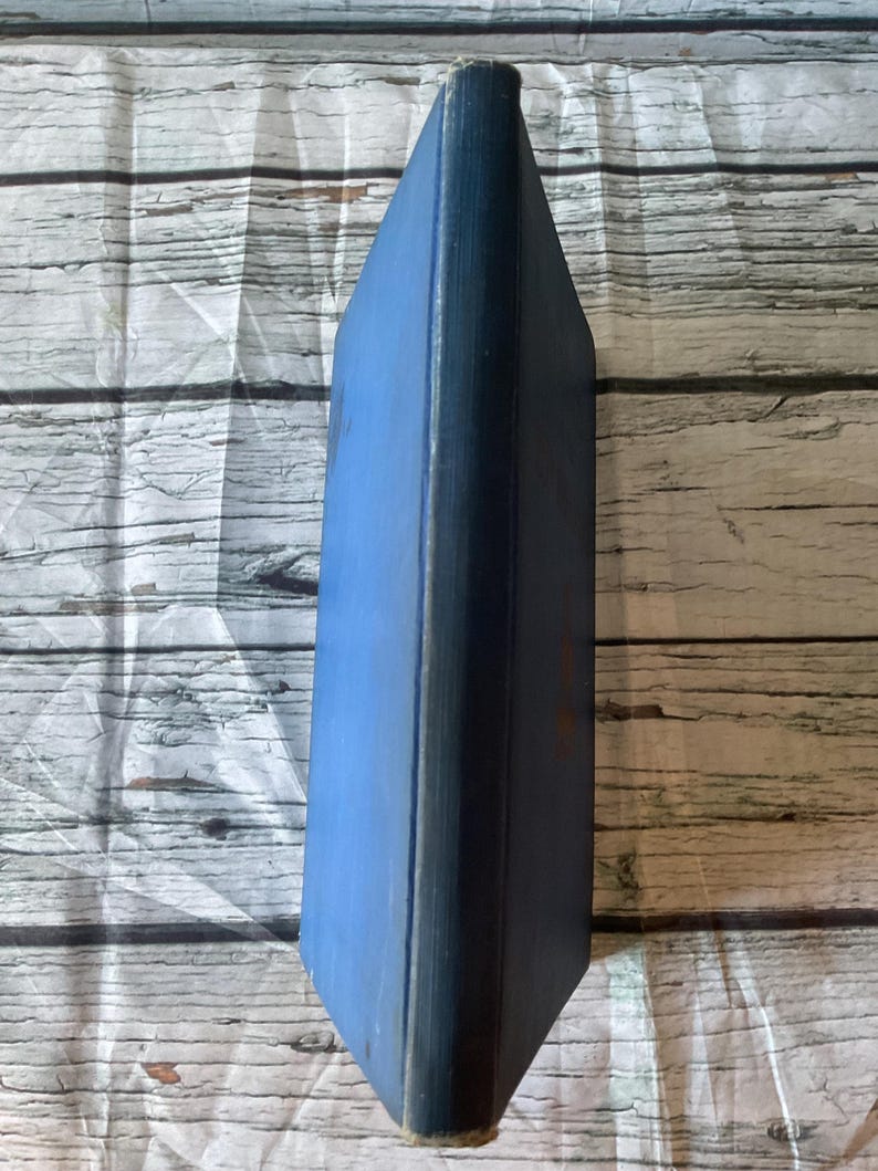 May include: A vintage book with a blue cover and spine. The book's edges are worn, showing signs of age. The cover is a solid blue, while the spine is a darker shade. The book is set against a light-colored, wood-grain background.