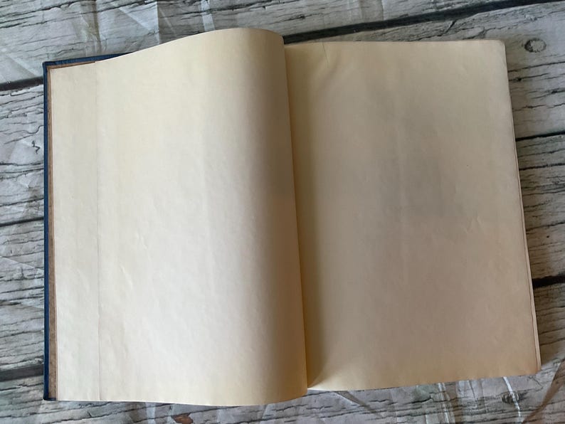 May include: An open book with aged, cream-colored pages. The book's spine is dark blue, and the pages are slightly yellowed, suggesting age. The book is lying open on a weathered, light-colored wooden surface.