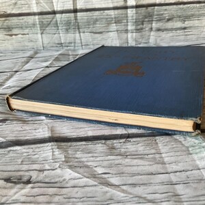May include: A vintage hardcover book with a blue cover and gold lettering. The book's pages are aged, and the edges show wear. The book is lying on a white and gray patterned surface.