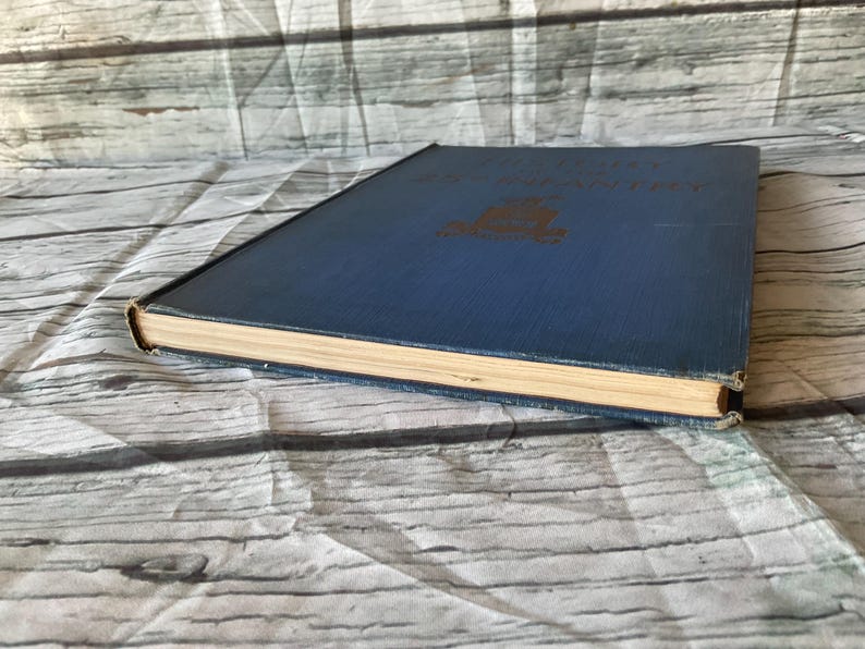 May include: A vintage hardcover book with a blue cover and gold lettering. The book's pages are aged, and the edges show wear. The book is lying on a white and gray patterned surface.