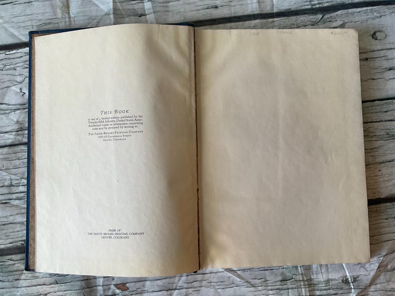 May include: Open book with aged, cream-colored pages. The left page has text that reads "THIS BOOK" and additional text below. The book's spine is dark blue. The right page is blank. The book is open on a light wood surface.