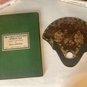 Op de afbeelding: Een antieke waaier met een sepia-afbeelding van drie kinderen, naast een groen boek getiteld "The Green Pastures Spirituals" van Hall Johnson. De waaier heeft een decoratieve rand met bloemen- en vlindermotieven.