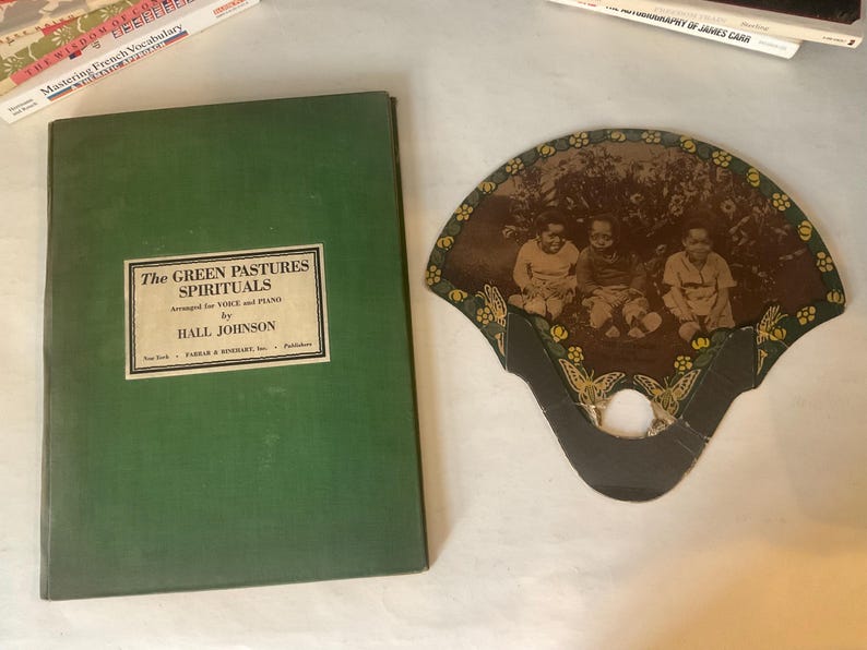 Op de afbeelding: Een antieke waaier met een sepia-afbeelding van drie kinderen, naast een groen boek getiteld "The Green Pastures Spirituals" van Hall Johnson. De waaier heeft een decoratieve rand met bloemen- en vlindermotieven.