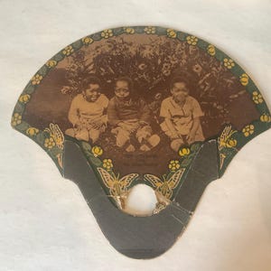 Op de afbeelding: Antieke waaier met een sepiafoto van drie kinderen. De waaier heeft een decoratieve rand met bloemen- en vlindermotieven. De handgreep is donkergrijs en de bovenrand heeft een groene rand met gele bloemen.