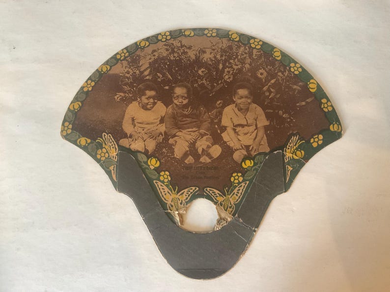 Op de afbeelding: Antieke waaier met een sepiafoto van drie kinderen. De waaier heeft een decoratieve rand met bloemen- en vlindermotieven. De handgreep is donkergrijs en de bovenrand heeft een groene rand met gele bloemen.