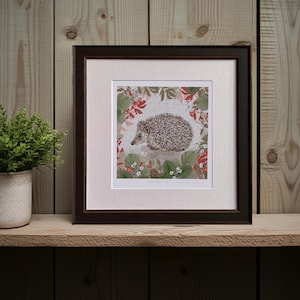 Peut inclure: Impression encadrée représentant un hérisson entouré de fraises rouges et de feuilles vertes. L'illustration est encadrée dans un cadre marron foncé et présentée sur une étagère en bois. Le fond est neutre.