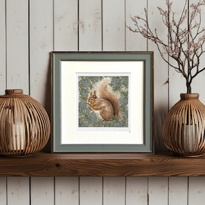 May include: Framed artwork featuring a detailed watercolor painting of a squirrel. The squirrel is depicted in shades of brown and tan, set against a backdrop of green foliage and blue pinecones. The artwork is framed in a muted green frame.