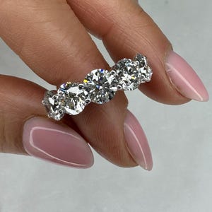 May include: A silver-toned ring featuring five round, brilliant-cut clear stones set in a row. The stones are held in place by metal prongs, creating a continuous band design that showcases the faceted surfaces and reflective quality of the stones.